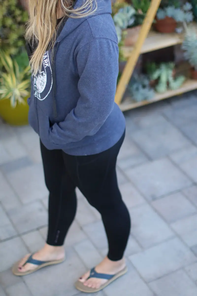 Woman standing on gray pavers outside wearing blue sandals, black leggings, and a blue zip up sweatshirt
