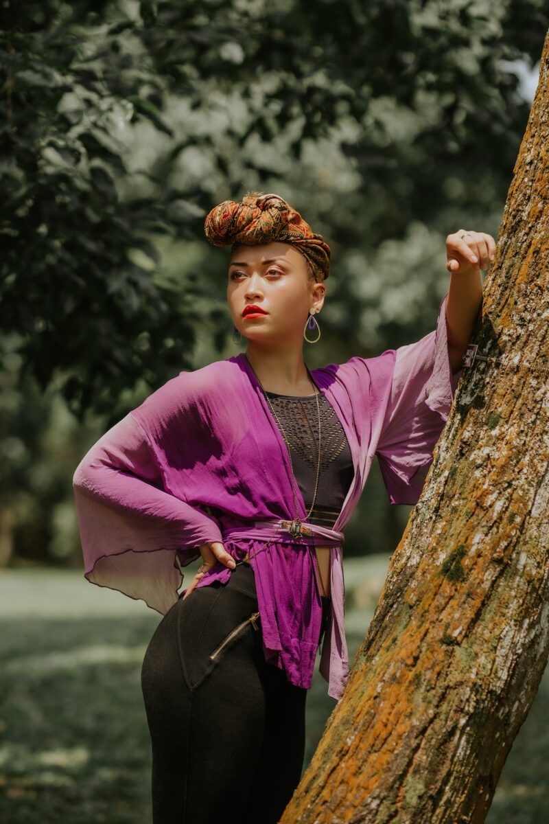 A woman in a black top and a purple flowy blouse wearing black leggings standing in front of a tree