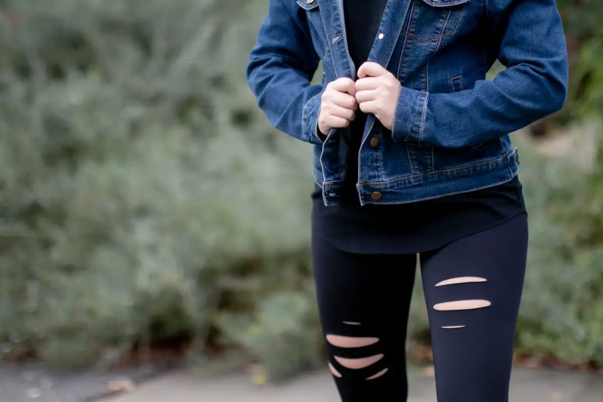 Woman wearing a blue jean jacket and holding the sides of it while wearing black ripped leggings outside in the driveway