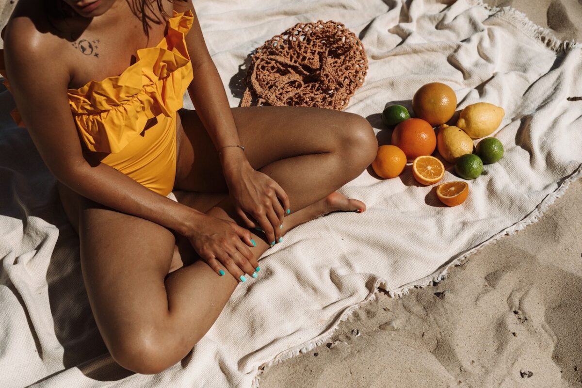 A woman with blue colored nail polish is wearing a yellow swimsuit with ruffles while sitting on a white cloth