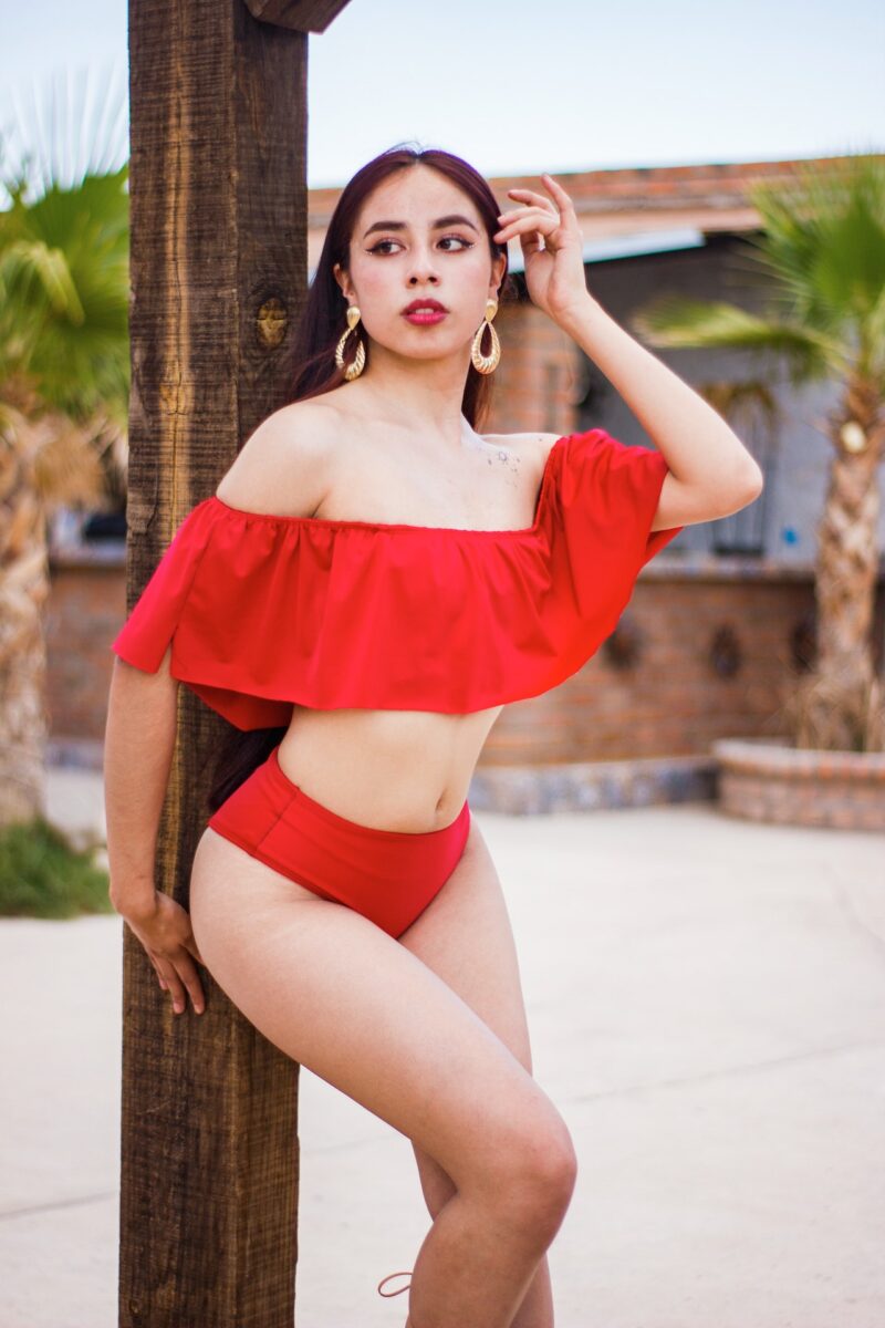 A woman wearing a red swimsuit and gold earrings is leaning on a brown wooden pole