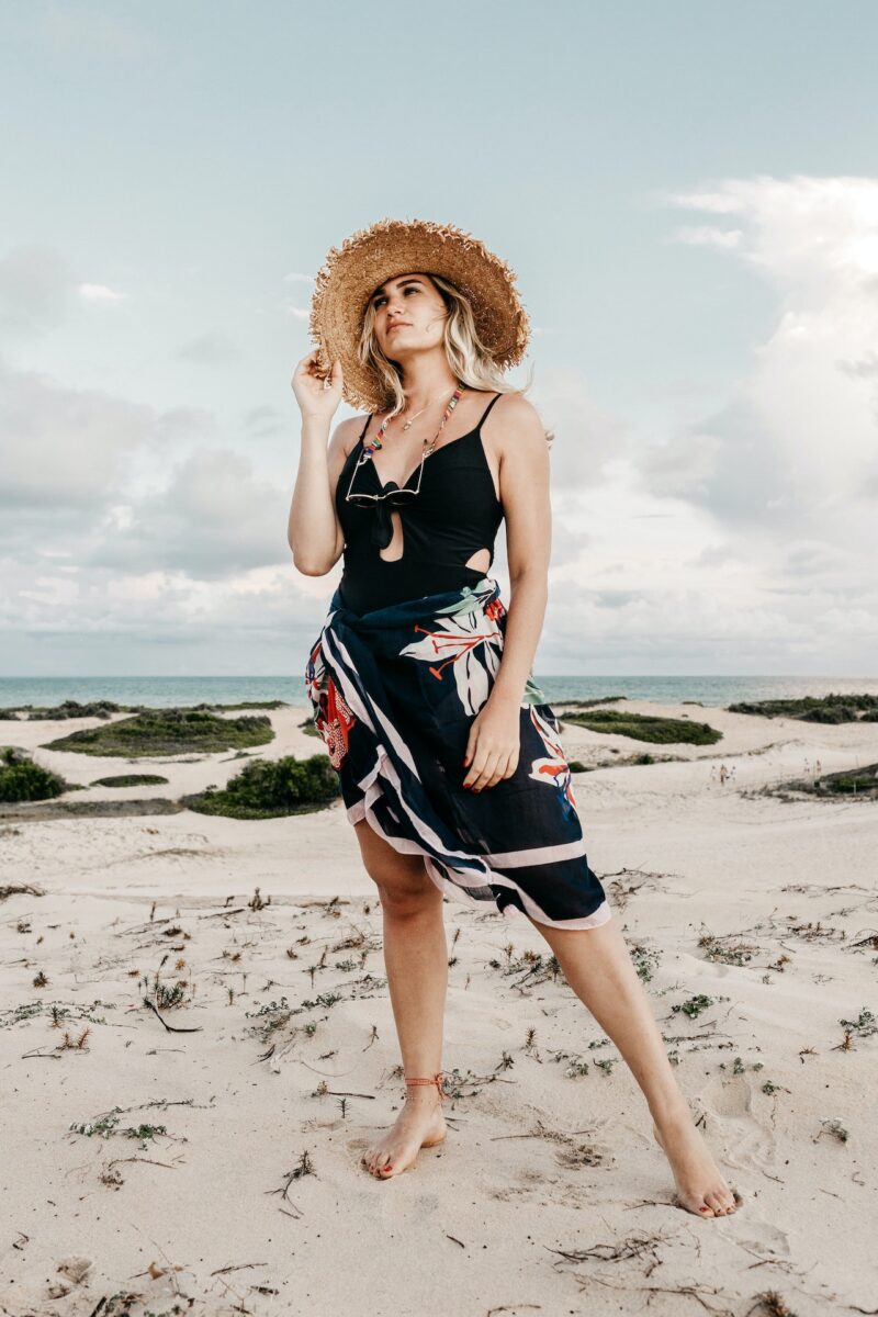 A woman wearing a black swimsuit and floral sarong, and brown sun hat while standing near the shore