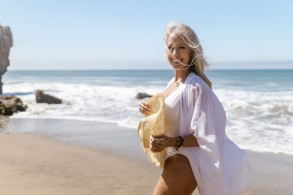 A woman wearing a white swimsuit and white coverup is holding a yellow sun hat while standing on the shore