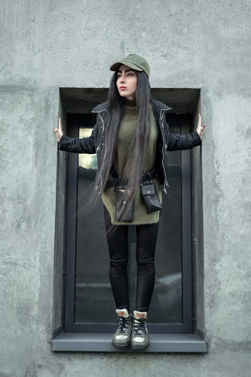 A woman in a black leather jacket and black leggings wearing a rubber shoes standing on a window sill