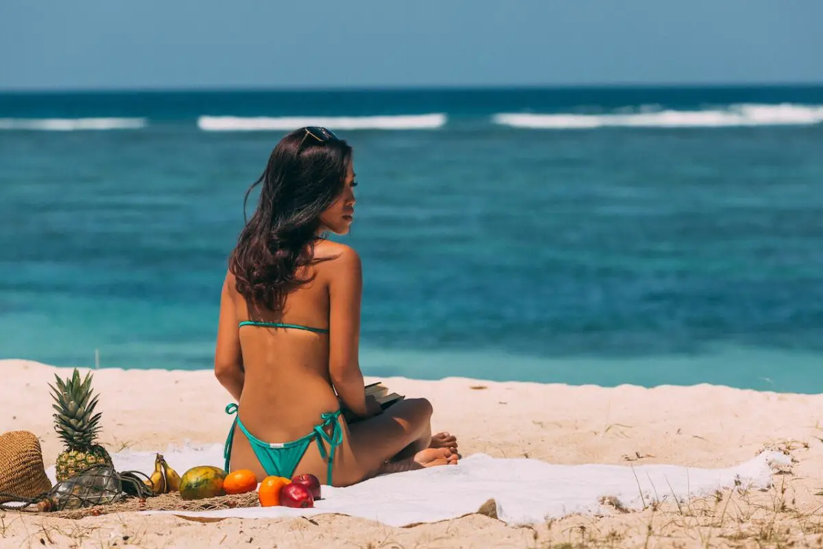A woman with long hair is wearing turquoise string bikini bottoms while sitting on a white beach towel with fresh fruits near the shore