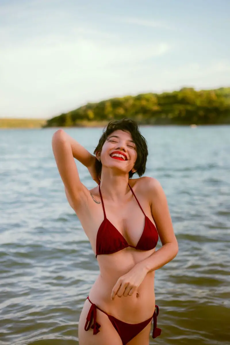 A woman with short hair is wearing a maroon string bikini and bikini top while standing near the calm beach
