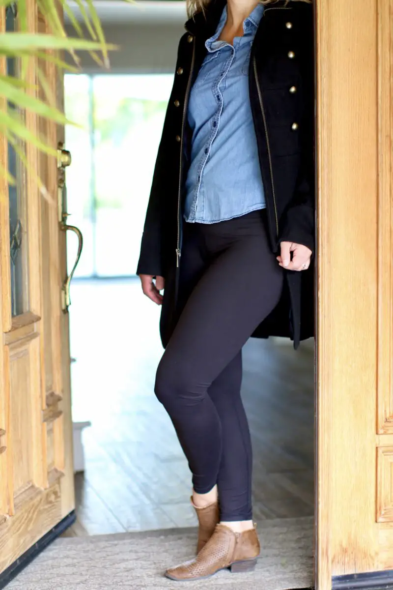 A woman with blonde hair is wearing a denim jacket, black leggings, and brown shoes while standing near the brown wooden door