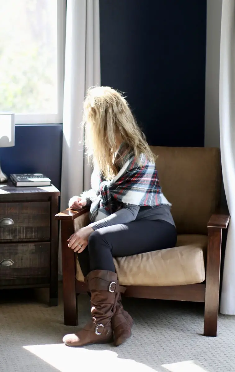 A woman with blonde hair is wearing a gray full sleeves tee, a checkered blanket scarf, brown suede boots, and leggings while sitting near a gray wall