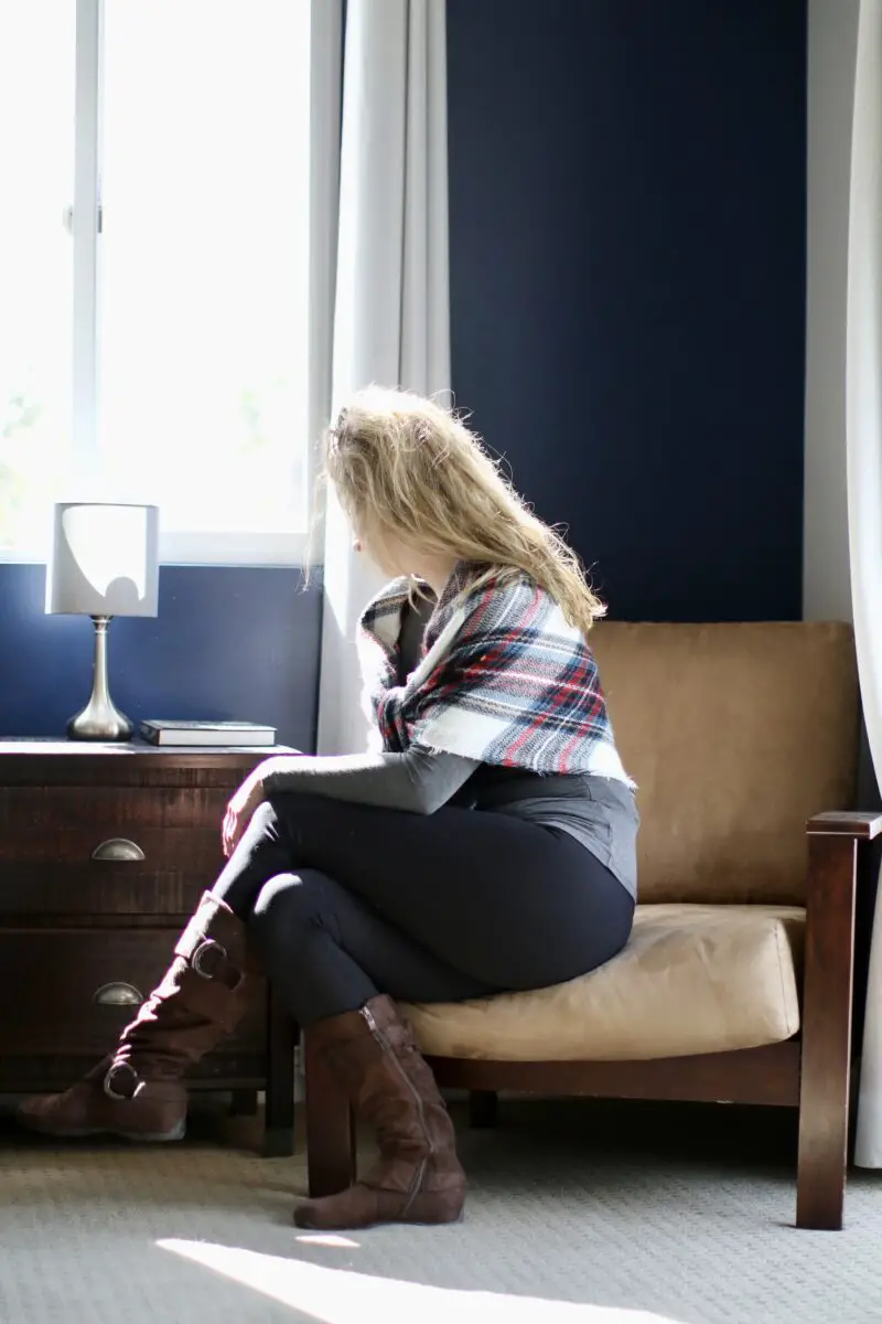 A woman with blonde hair is wearing a gray full sleeves tee, a checkered blanket scarf, brown suede boots, and leggings while sitting on a brown chair