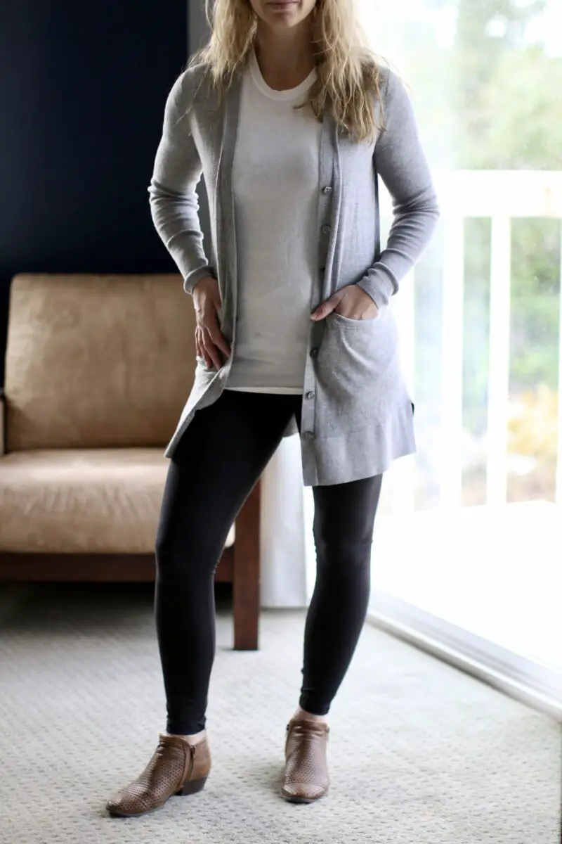 A woman wearing a long gray cardigan, white tee, brown ankle boots, and black leggings while standing on a gray carpet floor