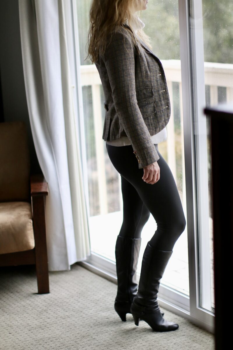 A woman wearing a long Button-Down, sweater vest, leggings, and knee-high boots while standing near a sliding glass door