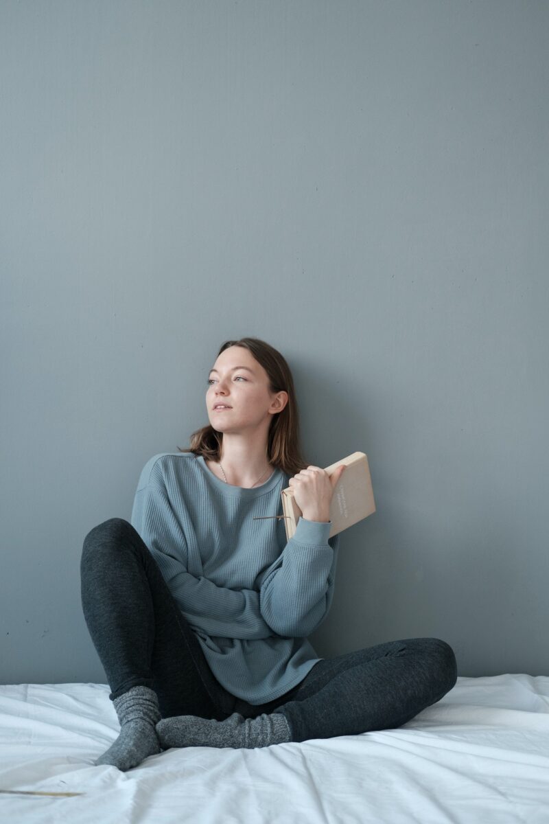A woman in a blue sweatshirt and black leggings holds a book while seated on the bed