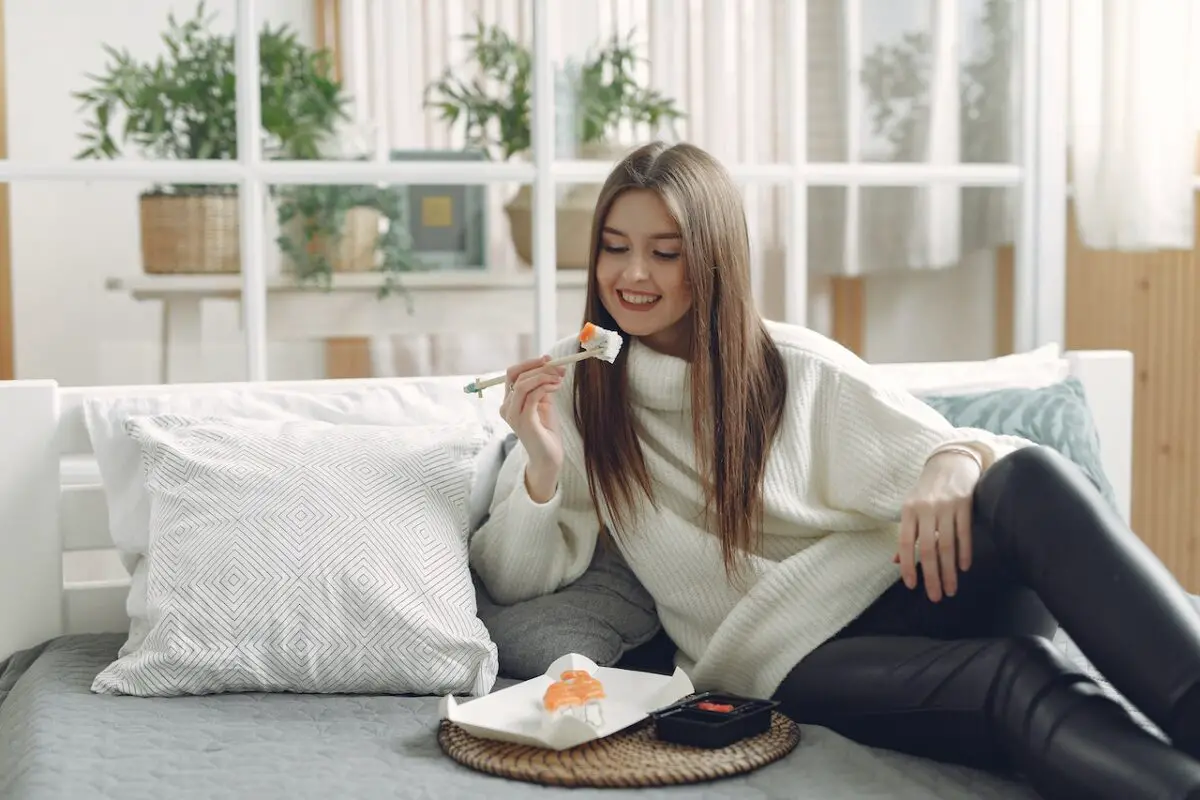 A young woman wearing a white sweatshirt and black leggings eating sushi at home