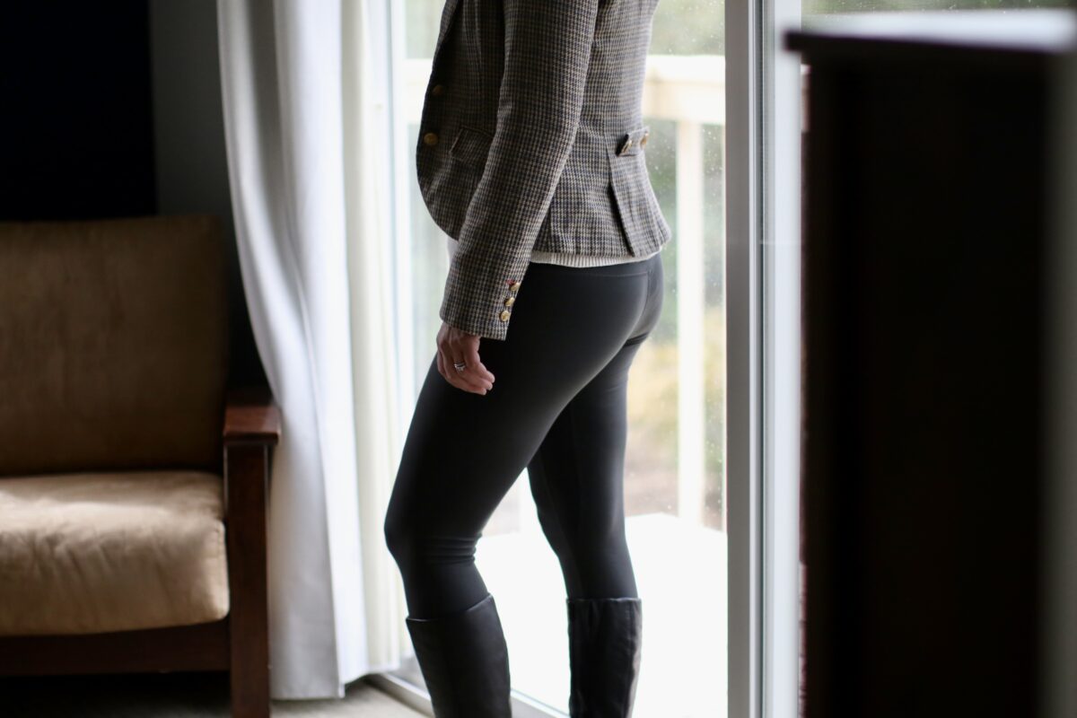 A person wearing a gray modern blazer, black leggings, and black high heel boots while standing near a glass sliding door