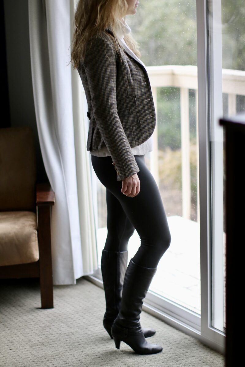 A woman with blonde hair wearing a gray modern blazer on top of a white turtle neck sweater, black leggings, and black high heel boots while looking outside the sliding door