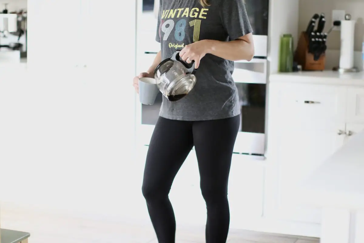 A person wearing a gray graphic shirt and black leggings while holding a gray mug and coffee pot