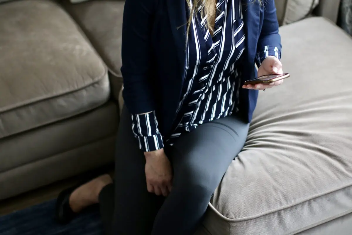 A woman wearing a black and white button-down shirt, black leggings, and brown flats is sitting on a gray sofa while holding a smartphone