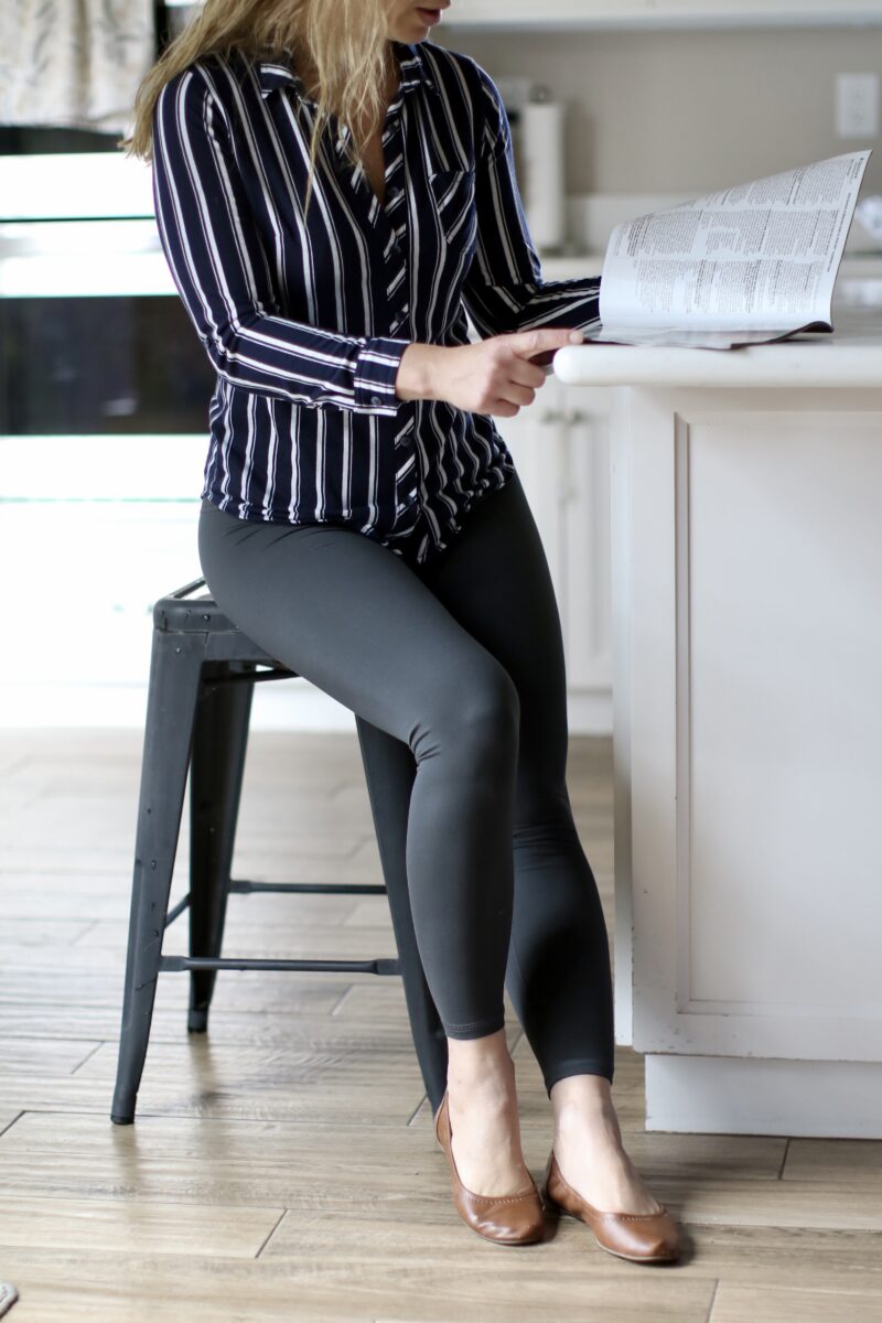 A woman wearing a black and white button-down shirt, black leggings, and brown flats is sitting on a black chair