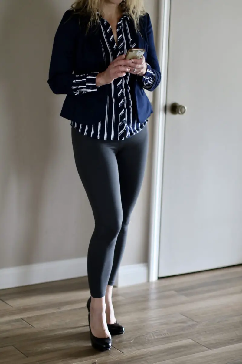 A woman wearing a black and white button-down shirt, leggings, and brown flats is standing on a brown wooden floor while holding a smartphone