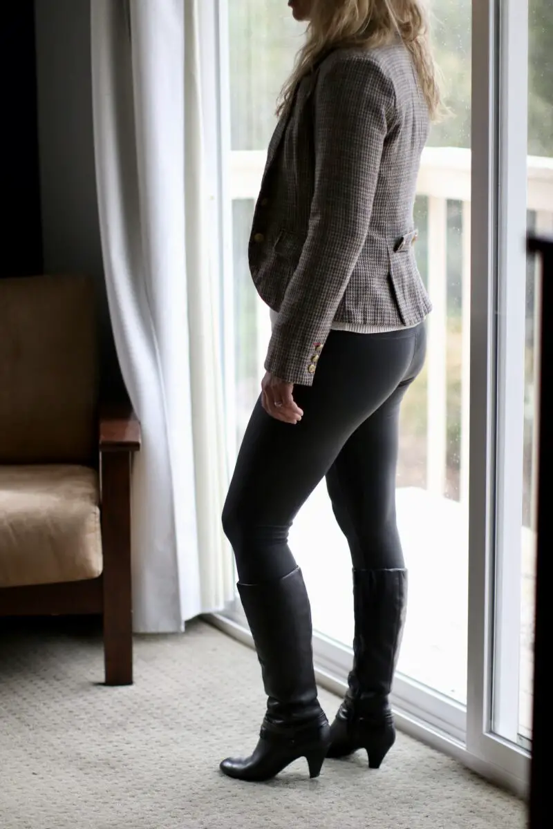 A woman wearing a modern blazer, leggings, and high heel boots while standing near a glass sliding door