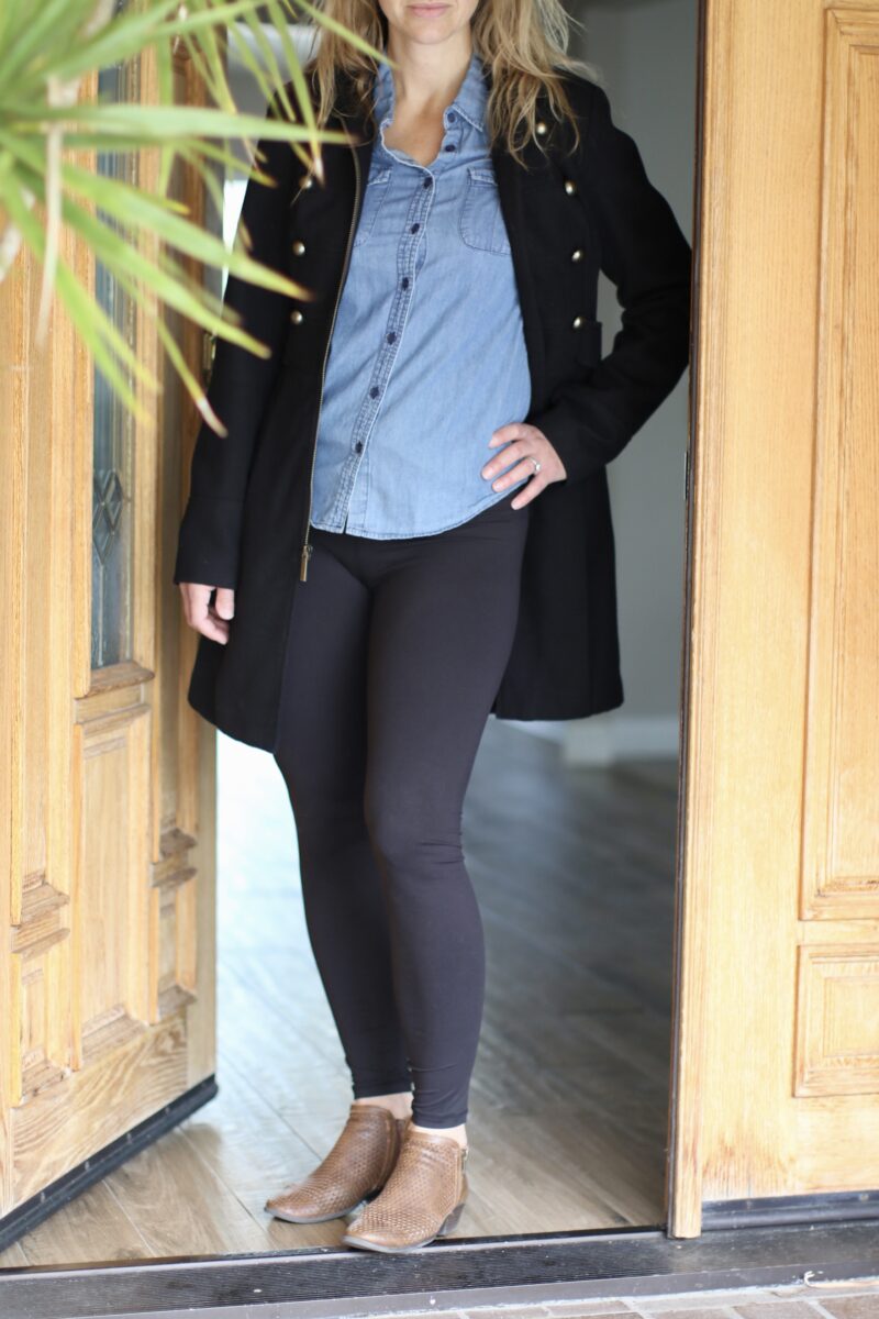 A woman wearing a denim jacket, black leggings, and brown shoes is standing near the brown wooden door