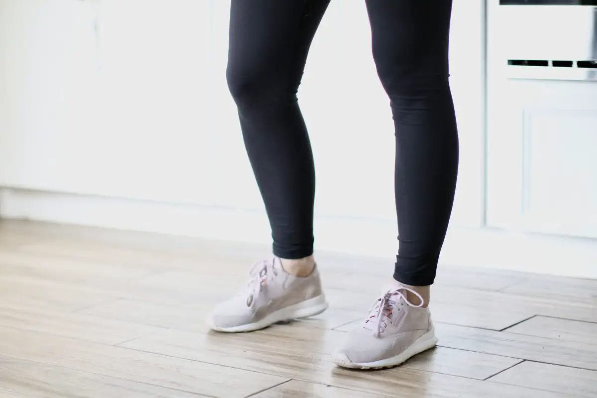 A person wearing black leggings and pink sneakers is standing on a brown wooden floor