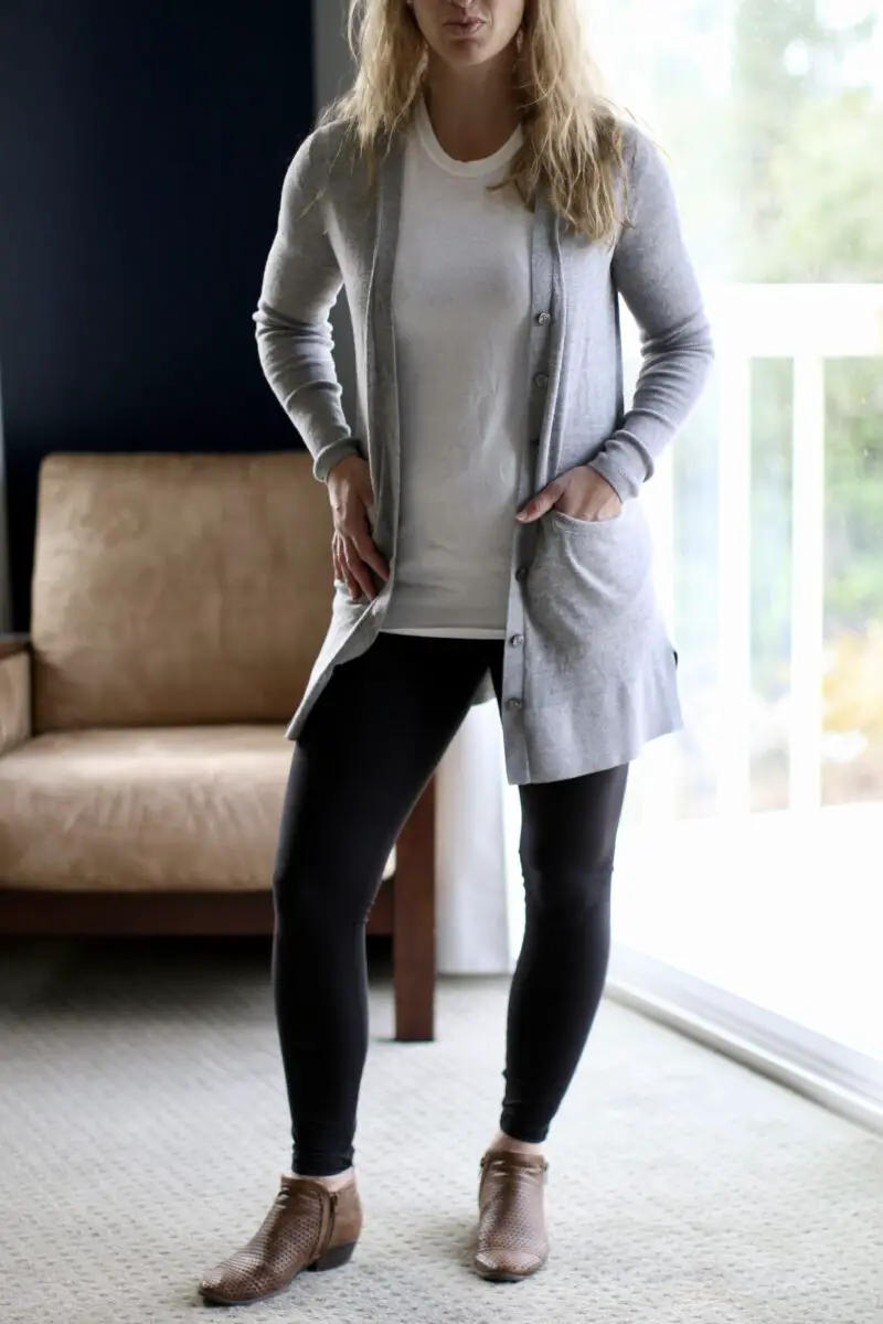 A woman wearing a gray cardigan on top of a white tee, black leggings, and brown ankle boots while standing on a gray-carpeted floor