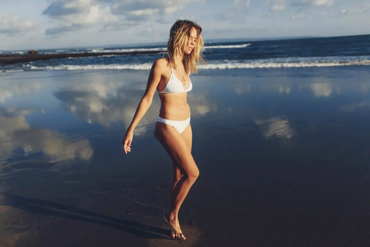 A young woman wearing a white bikini walking on a beach against a cloudy sky reflecting in the water