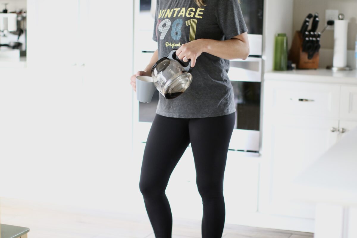 A person with blonde hair is wearing a gray graphic shirt and black leggings while holding a gray mug and coffee pot