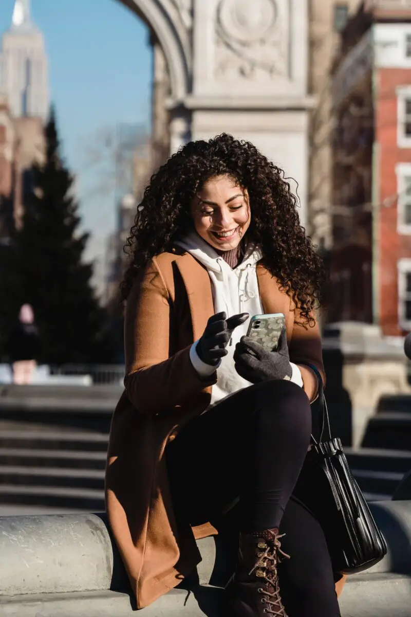 A woman with long curly hair is wearing a brown coat and black leggings while using her smartphone outdoors