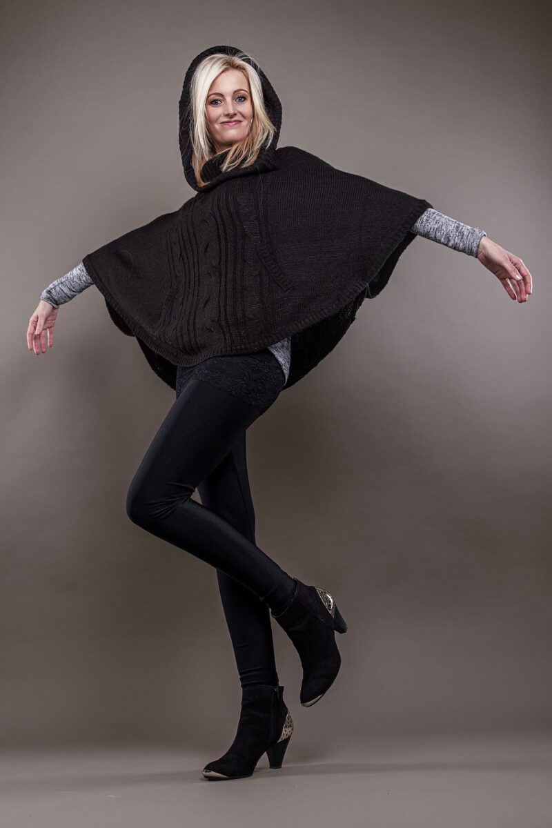 A woman with blonde hair is wearing a black poncho on top of a gray long sleeves shirt and black leggings while standing on a gray background