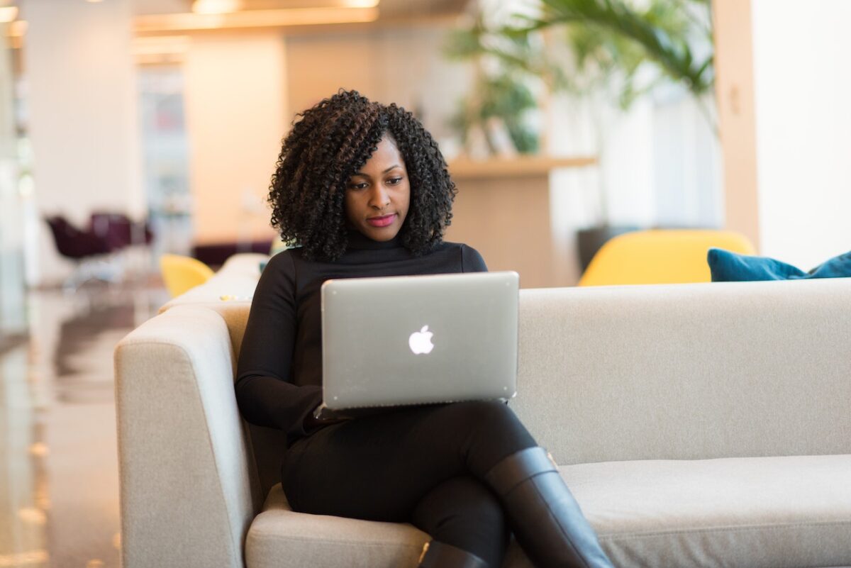 A woman using a silver laptop is wearing a black turtle neck, black leggings, and boots while sitting on a gray couch