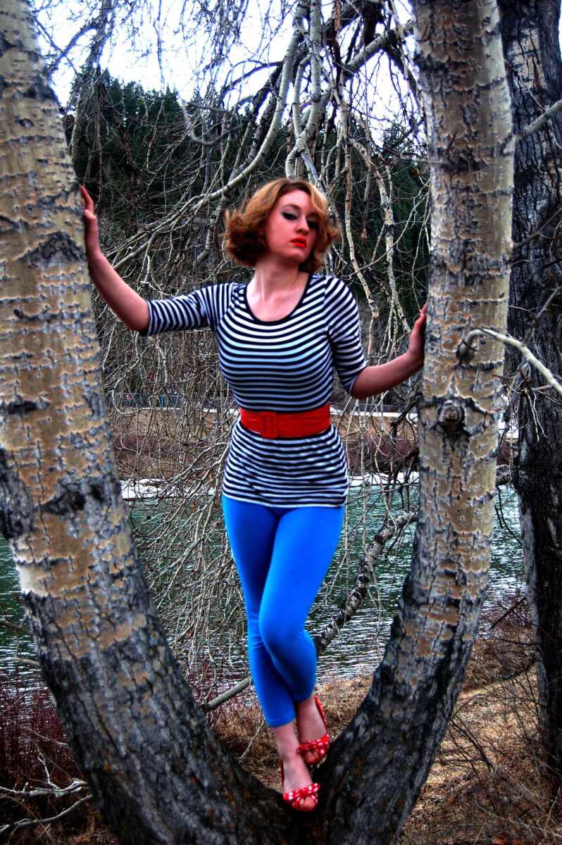 Woman wearing a long-sleeved black and white striped shirt with a red belt and blue stockings while standing on a tree branch