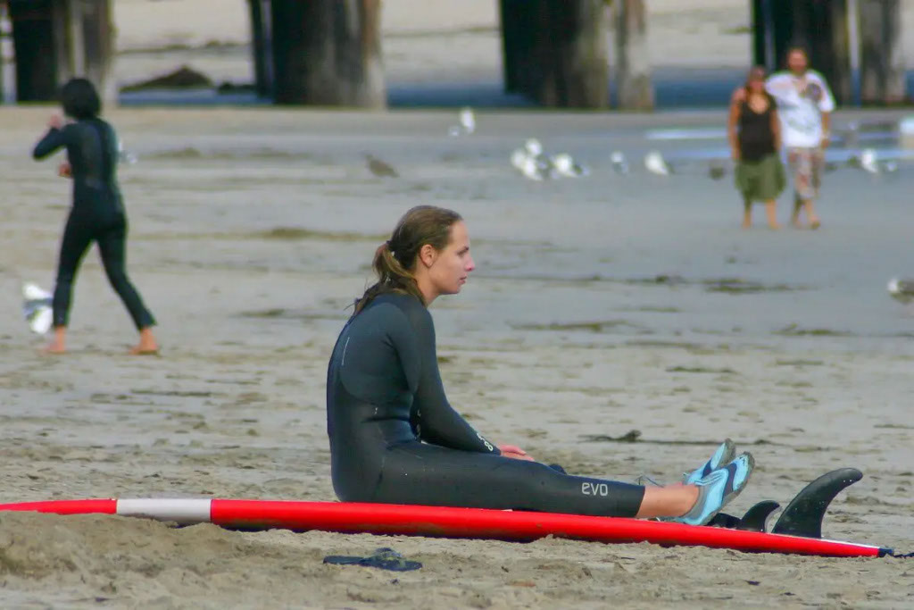 Woman wearing a black wetsuit while sitting on top of her red surfboard while observing the sea water level