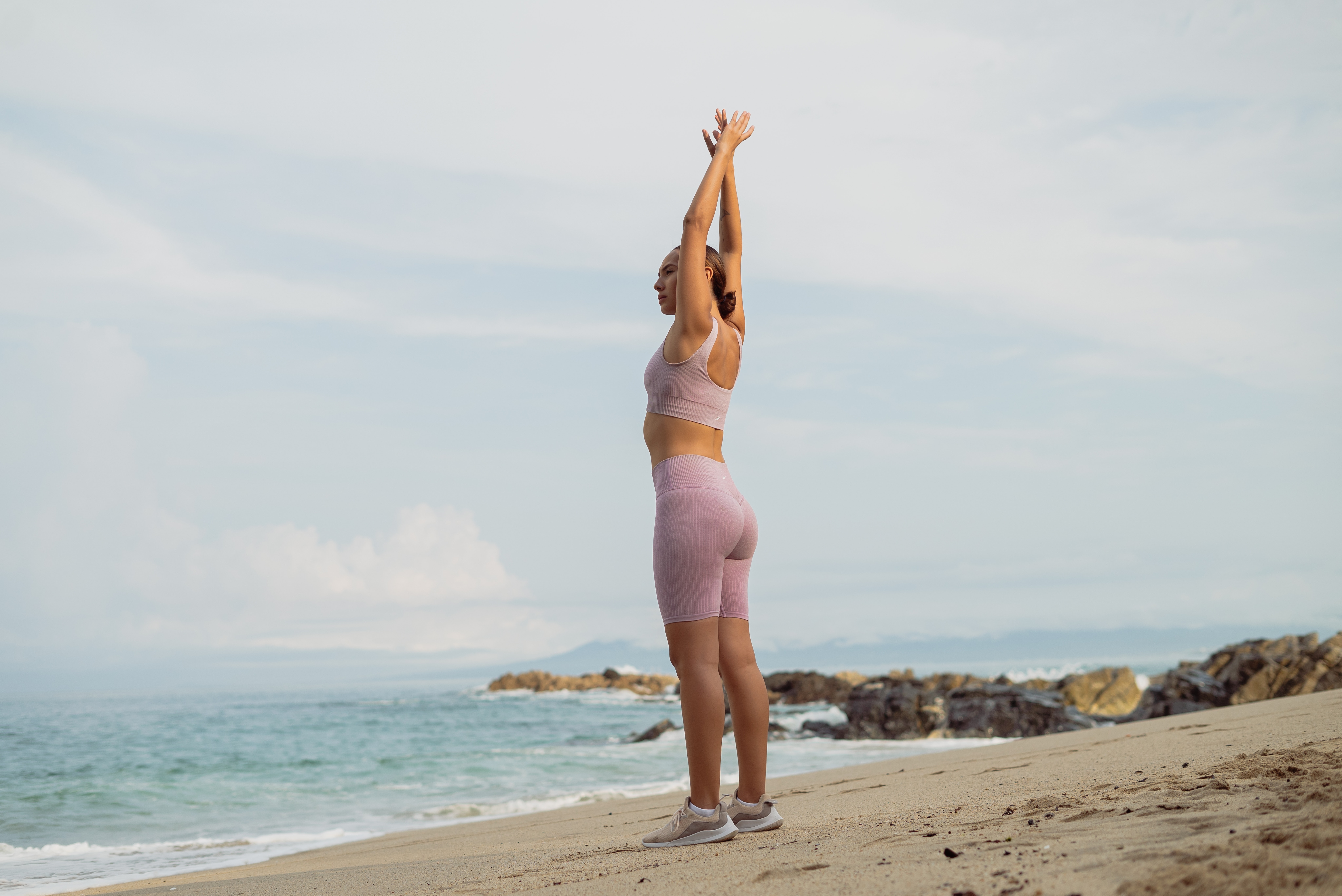A woman who is stretching wearing sports bra, shorts, and shoes at the seashore