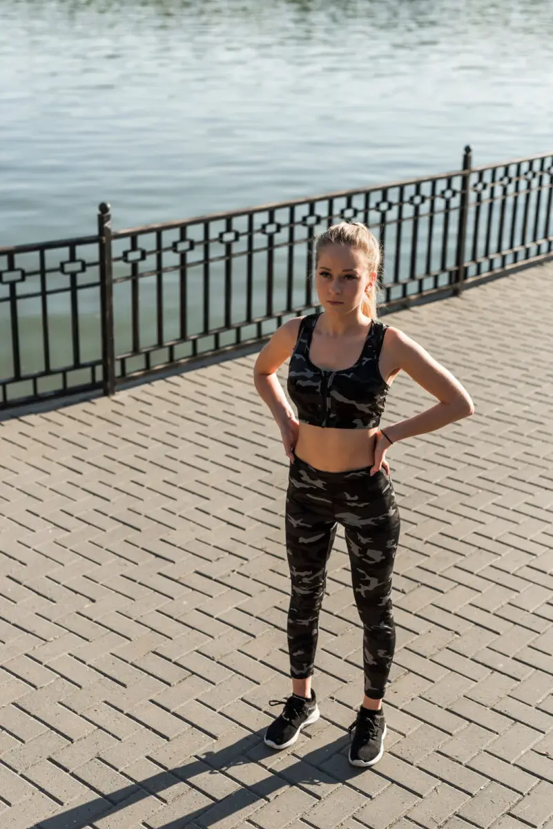 A woman wearing an army green camo leggings and sports bra standing in a park near a river