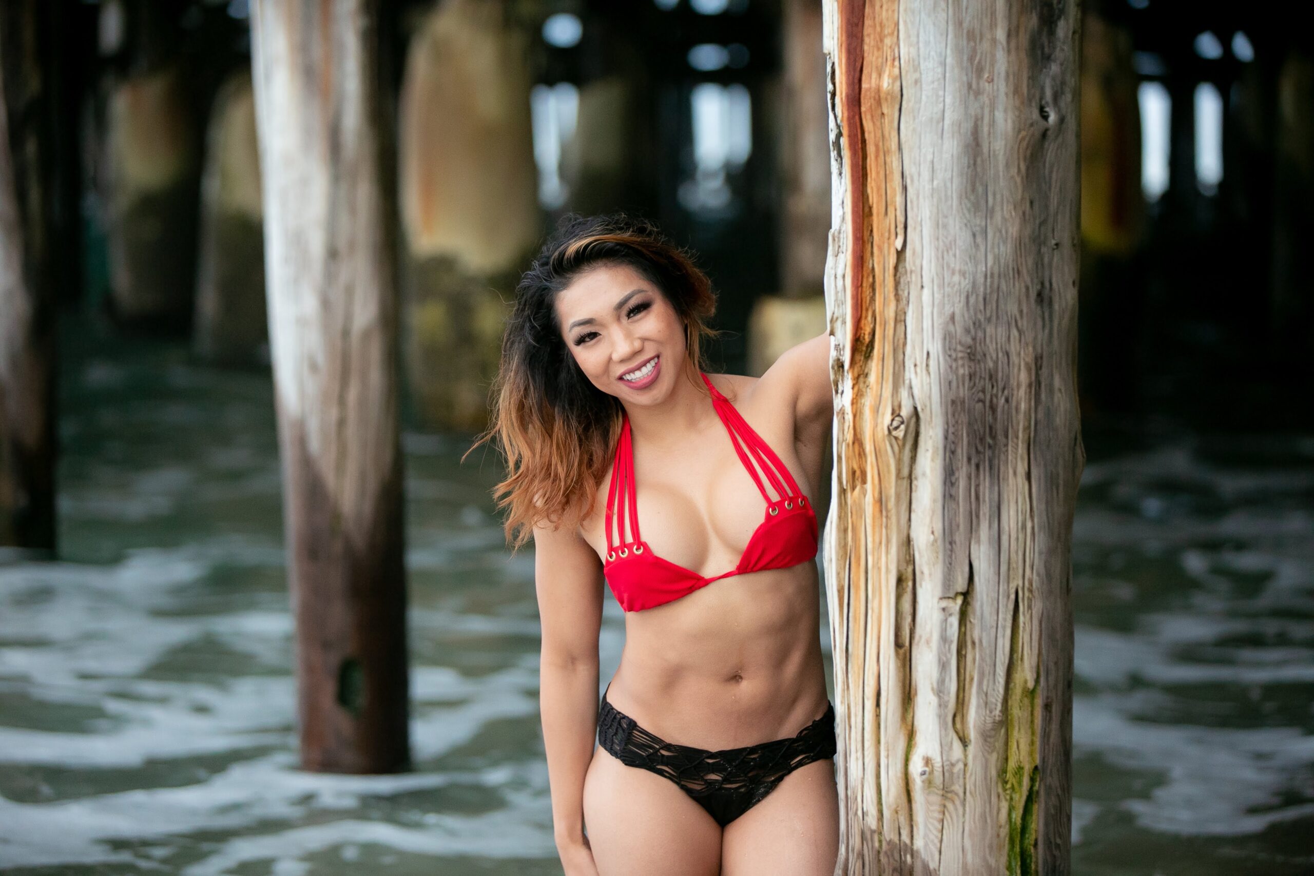 Woman wearing a two-piece leaning a thick brown wood below a wooden beach bridge