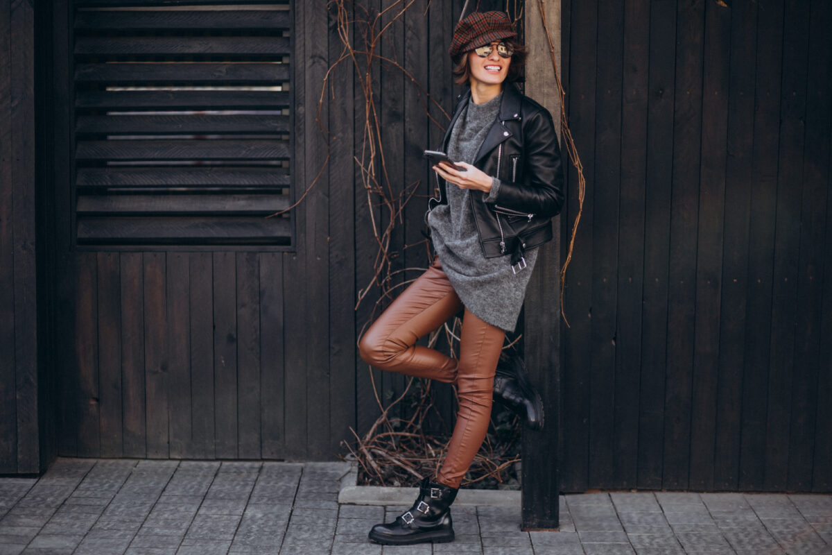 A woman wearing a black jacket, sweater and brown leggings leaning on a wooden post holding a phone